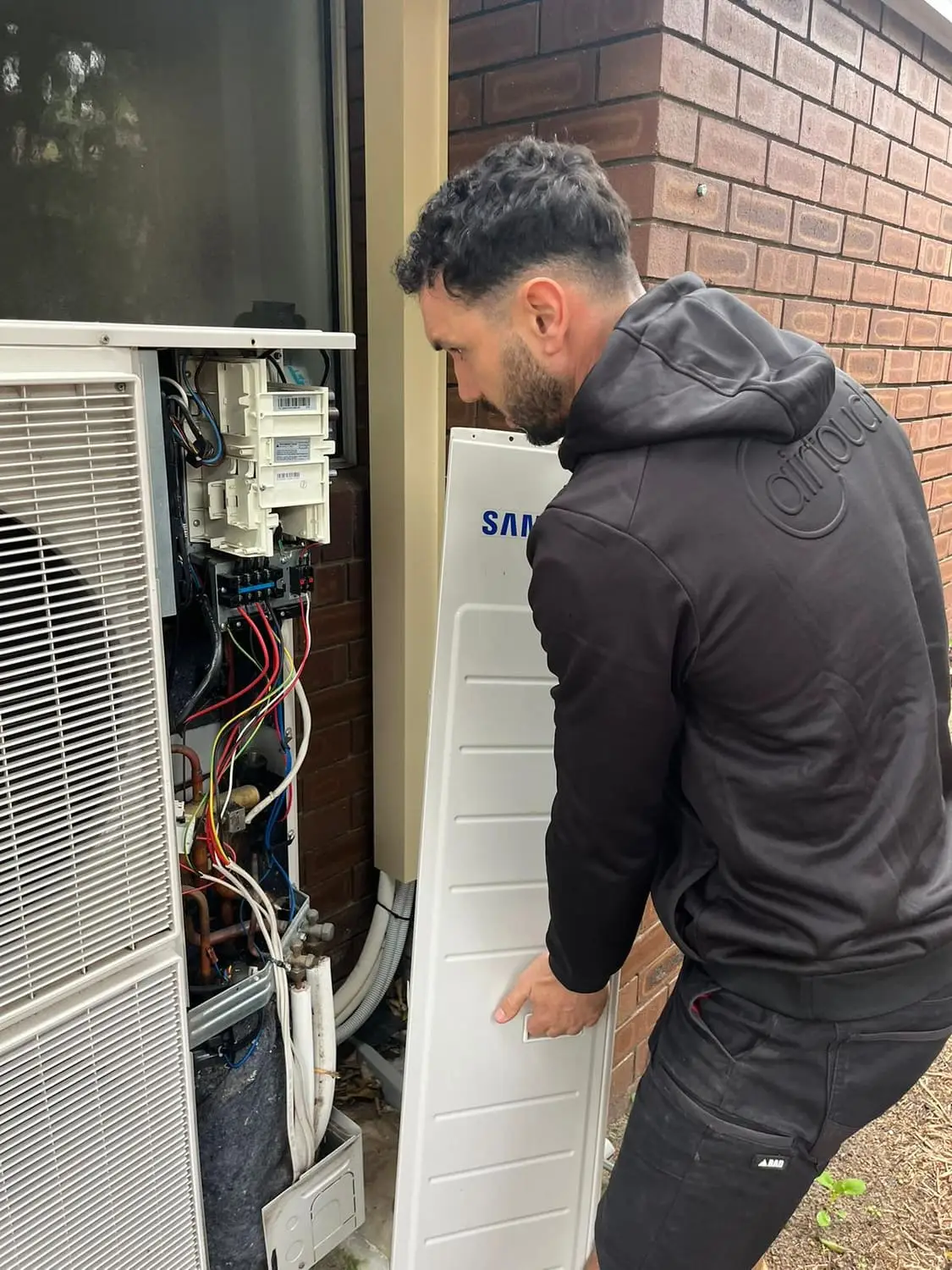 Aerobreeze technician working inside an opened outdoor condensing unit against a brick wall