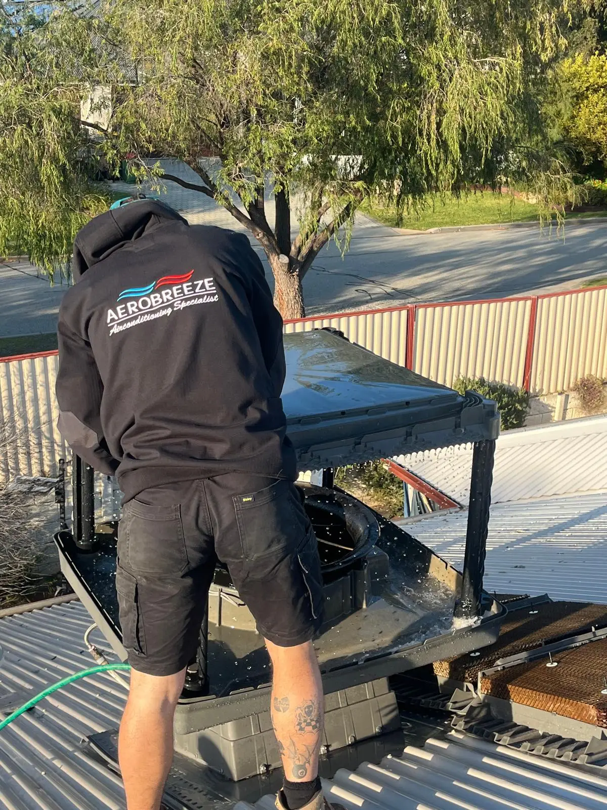 Aerobreeze technician in uniform flushing out an opened evaporative unit on a Colorbond roof, the pads lifted clear